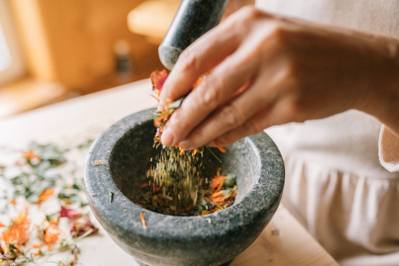 Hands grinding colorful dried herbs and petals in a stone mortar on a wooden table.